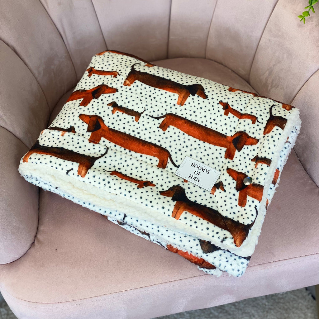 Blanket with dachshund pattern on a beige chair, featuring 'Hounds of Eden' branding.
